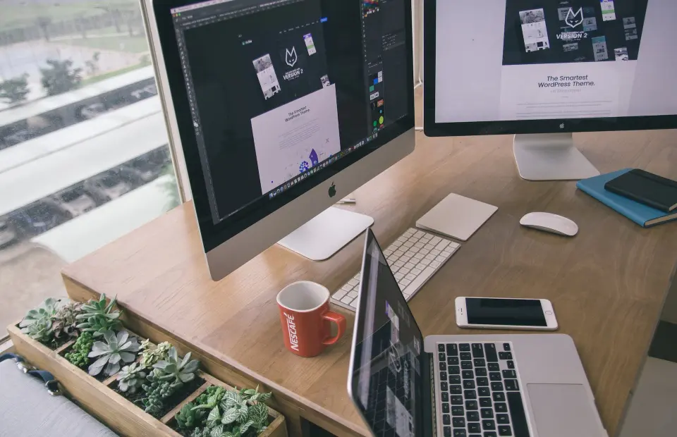 A PRO consultant's work desk is equipped with two computer monitors, a laptop, a mobile phone, a notebook, and a coffee cup.